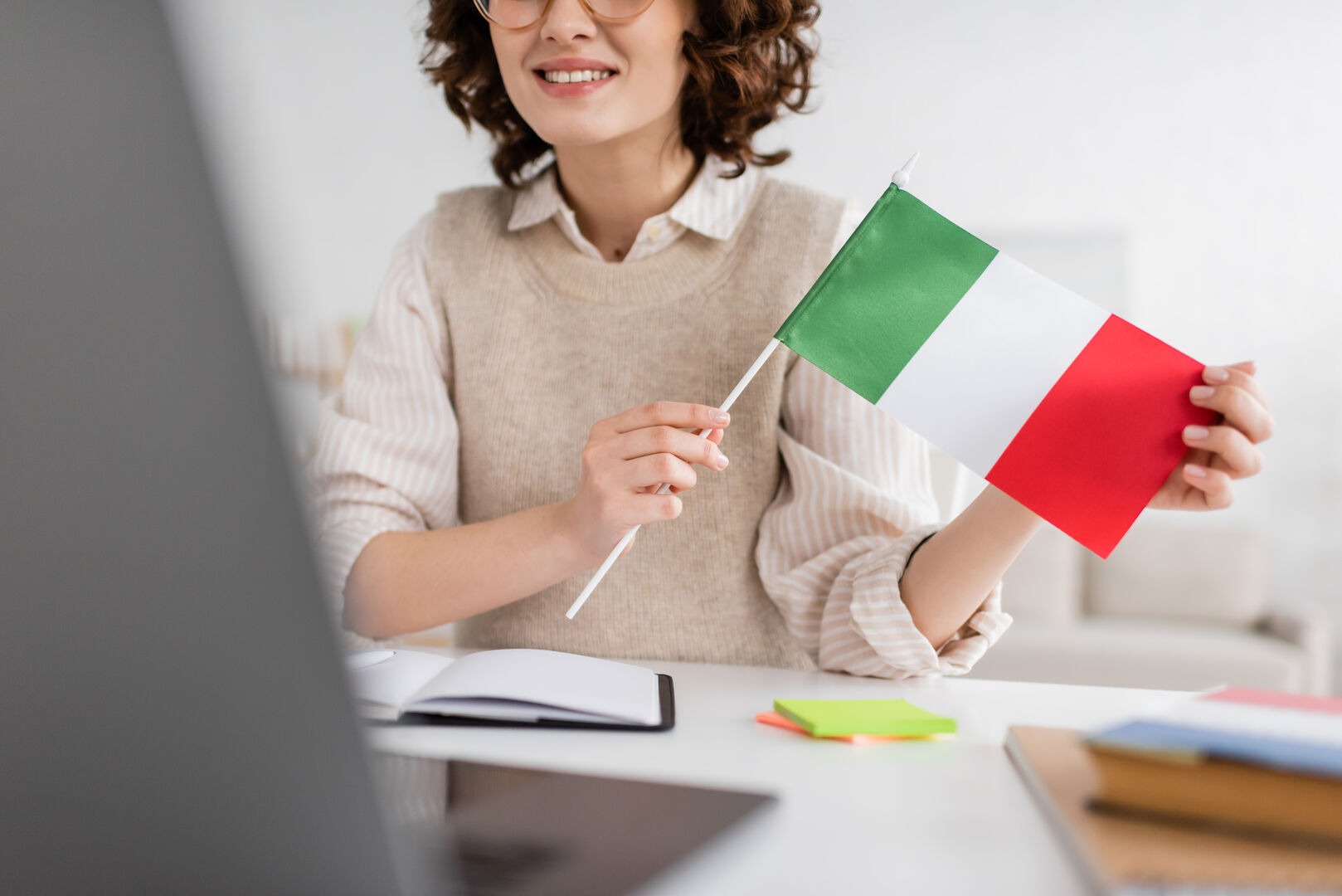 partial view of smiling language teacher holding flag of Italy during online lesson on blurred laptop at home